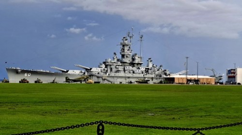 A large battleship docked at a harbor, surrounded by green grass and military vehicles under a cloudy sky.