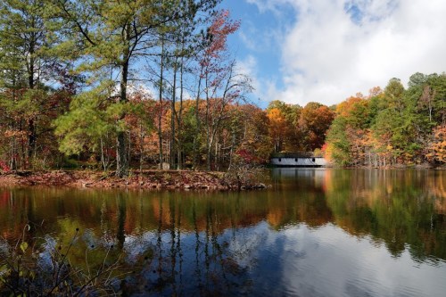 A serene lake surrounded by colorful autumn trees, reflecting the vibrant foliage and a cloudy blue sky.