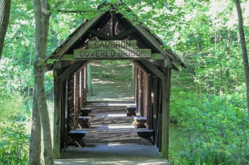 A wooden covered bridge surrounded by lush green trees, with a sign reading "Cambron Covered Bridge."