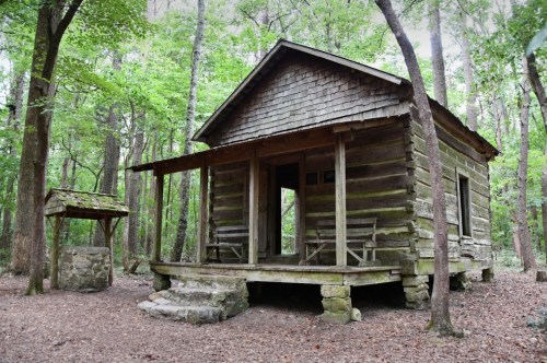 A rustic log cabin surrounded by trees, featuring a porch and a nearby stone well in a wooded area.