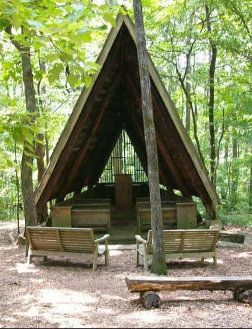 A wooden A-frame shelter surrounded by trees, featuring benches and a central altar in a serene forest setting.