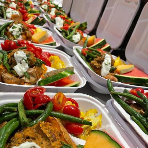 A row of takeout containers filled with colorful vegetables, tomatoes, and slices of fruit.