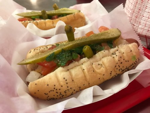 Two Chicago-style hot dogs topped with pickles, tomatoes, and green relish, served in a red tray with white paper.