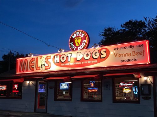 A brightly lit sign for Mel's Hot Dogs, featuring Chicago-style hot dogs and Vienna Beef products, at dusk.