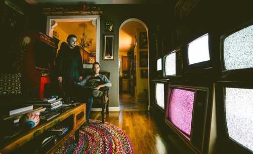 A person in a mask stands beside another seated in a cozy room filled with vintage TVs displaying static.