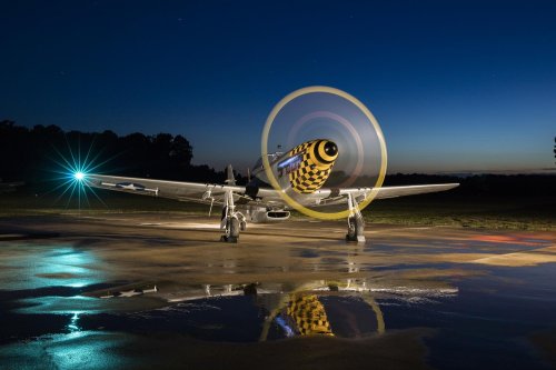 A vintage airplane with a colorful propeller, illuminated by a blue light, on a wet runway at dusk.