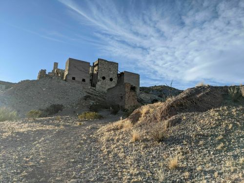 Ruins of a stone building on a hillside under a blue sky with scattered clouds. Desert landscape in the foreground.
