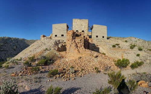 Abandoned stone buildings on a hillside, surrounded by rocky terrain and sparse vegetation under a clear blue sky.