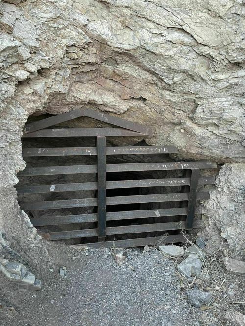 Entrance to a rocky mine shaft, partially covered with wooden slats and surrounded by rough terrain.