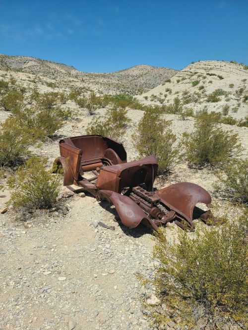 Rusty vintage car chassis abandoned in a desert landscape, surrounded by sparse vegetation and rocky hills.