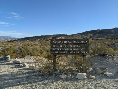 A weathered sign in a desert landscape warns of hazardous areas and advises to keep off structures due to mercury and open mine shafts.