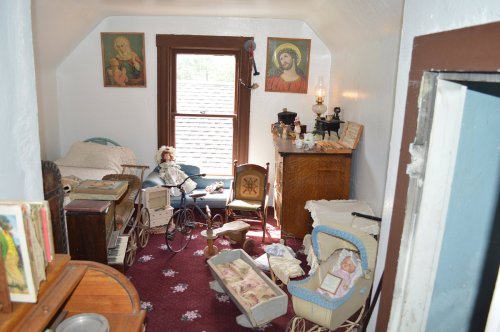 A vintage room with dolls, furniture, and religious artwork, featuring a bed, a rocking chair, and a window.