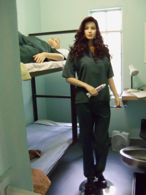 A mannequin in a prison uniform stands in a cell with a bunk bed and a sink, depicting a correctional facility setting.