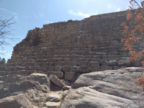 Ancient stone structure with large steps, surrounded by rocks and sparse trees under a clear blue sky.