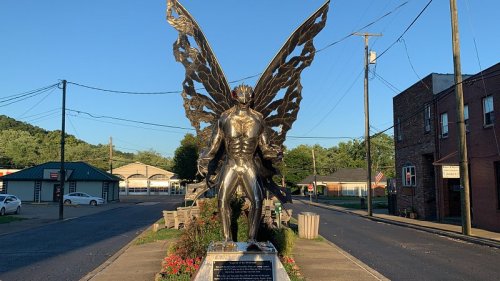 A large metallic statue of a figure with wings stands in a street, surrounded by buildings and greenery.