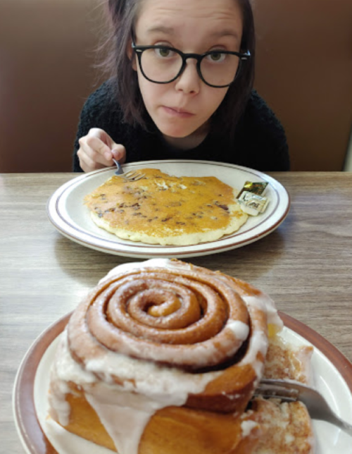 A girl with glasses looks at a plate of pancakes while a cinnamon roll sits in the foreground.
