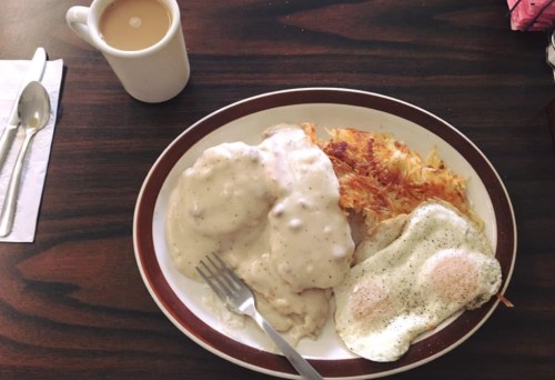 A plate of biscuits and gravy, hash browns, and two fried eggs, with a cup of coffee in the background.