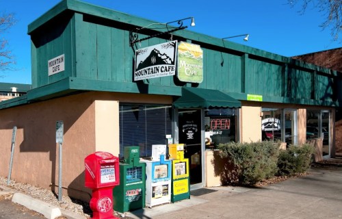A cozy café with a green awning, outdoor seating, and newspaper boxes in front, set against a clear blue sky.