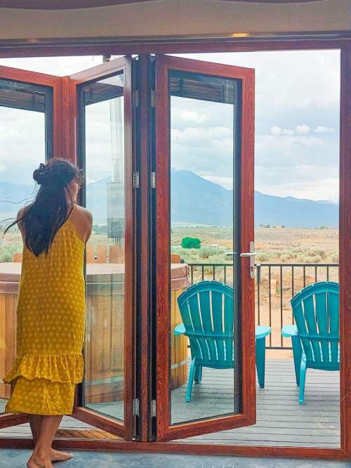 A woman in a yellow polka dot dress stands by open double doors, overlooking a scenic view with blue chairs outside.