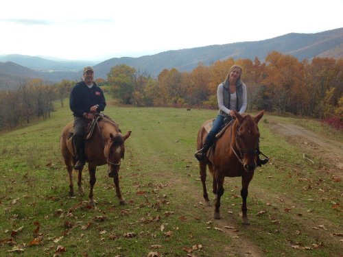 Two riders on horseback on a grassy trail, surrounded by autumn trees and mountains in the background.