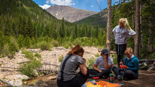A group of four people enjoying a meal outdoors by a river, surrounded by trees and mountains in a scenic landscape.