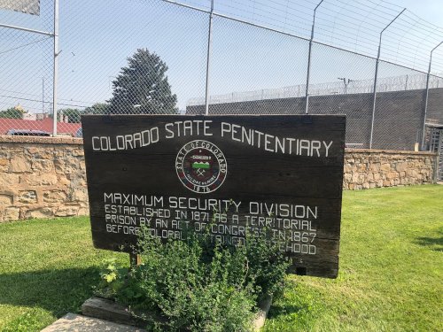 Sign for Colorado State Penitentiary, detailing its establishment and maximum security division. Fenced prison in background.