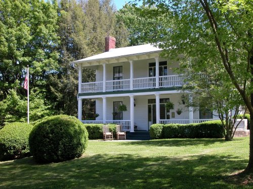 A two-story white house with a porch, surrounded by green trees and manicured bushes, featuring an American flag.