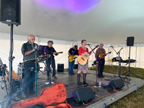 A band performs on stage under a tent, featuring a singer with an acoustic guitar and musicians with various instruments.