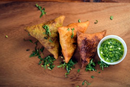 Three golden-brown samosas on a wooden surface, garnished with chopped herbs, alongside a small bowl of green chutney.