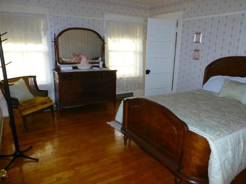 Cozy bedroom with a wooden bed, dresser, mirror, and a chair, featuring floral wallpaper and natural light.
