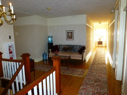 A hallway with a sofa, water cooler, and decorative rug, leading to a bright room at the end.
