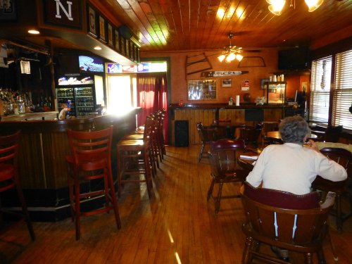 A cozy bar interior with wooden floors, empty chairs, and a person sitting at a table, enjoying the quiet atmosphere.