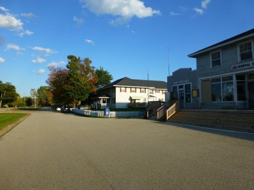 A quiet street scene featuring a general store and a house, with trees and a clear blue sky in the background.