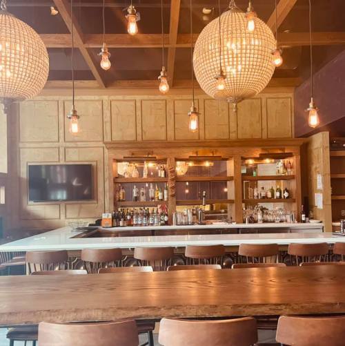 A cozy bar with wooden decor, pendant lights, and a well-stocked counter, featuring a large wooden table in the foreground.