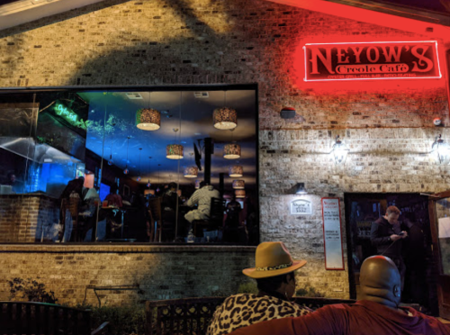 A lively night scene outside Neyow's Creole Café, with patrons visible through the windows and colorful lighting.