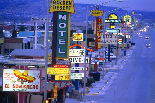 A colorful street scene featuring vintage motels and diners with neon signs along a highway at dusk.