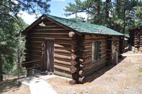 A rustic log cabin with a green roof, surrounded by trees and a dirt path leading to the door.
