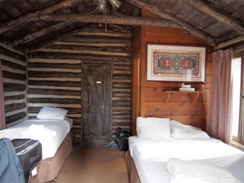 Cozy cabin interior with two beds, wooden walls, and a decorative wall hanging. Natural light from a window.