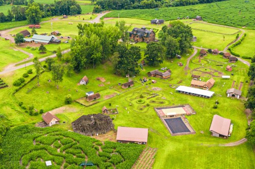 Aerial view of a green landscape with various buildings, fields, and pathways, surrounded by trees.