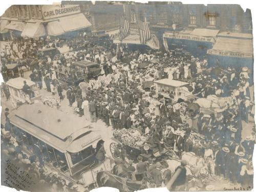 A bustling historical street scene with crowds, horse-drawn carriages, and early 20th-century attire.