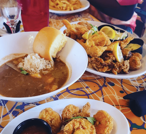 A plate of fried seafood with lemon, a bowl of gumbo with rice, and a side of bread on a colorful table.