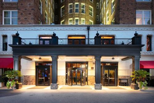 Elegant hotel entrance with large glass doors, decorative columns, and warm lighting, surrounded by potted plants.