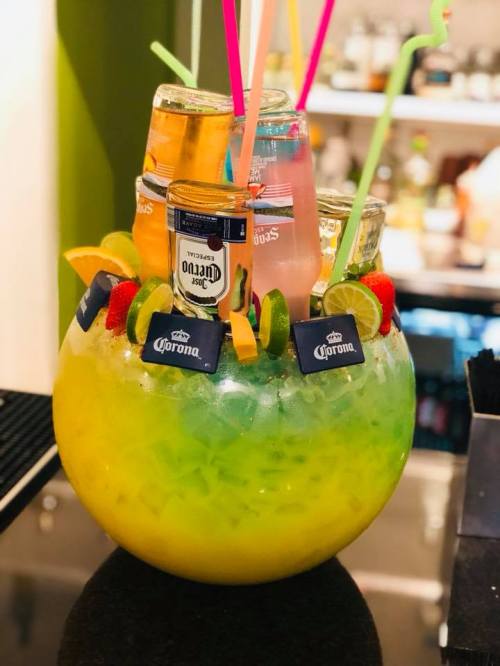 A colorful cocktail bowl filled with ice, fruit garnishes, and several drinks, including beer and colorful straws.