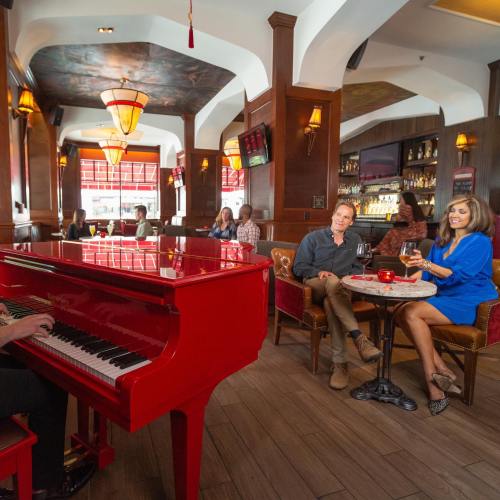 A pianist plays a red piano in a lively bar, with patrons enjoying drinks and conversation in a warm atmosphere.