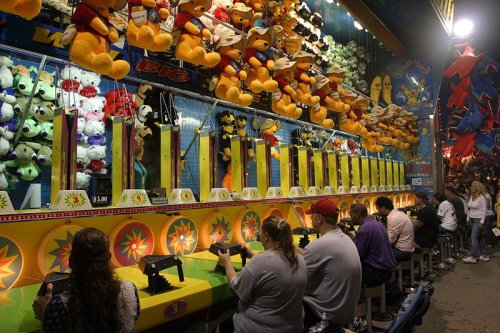 A carnival game booth with players aiming at targets, surrounded by colorful stuffed animals hanging above.