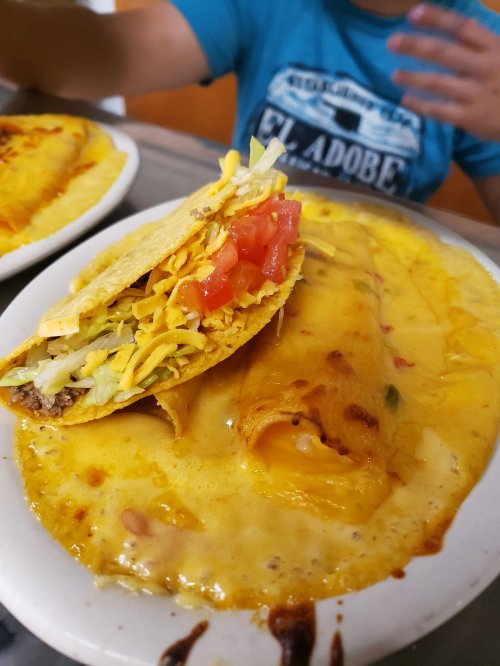 A plate featuring a cheesy enchilada and a taco filled with lettuce, cheese, and tomato, with a hand in the background.