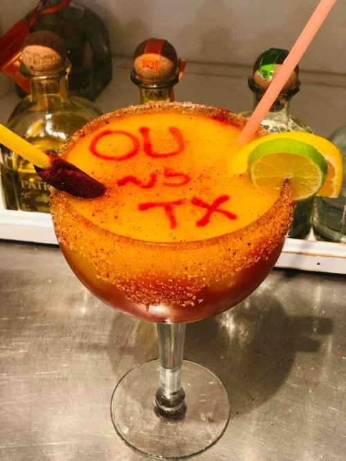 A colorful cocktail in a large glass, garnished with lime and a rim of chili powder, featuring "OU vs TX" written on top.