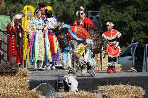 A group of Indigenous performers in traditional attire, showcasing a dance on a stage decorated with hay bales.