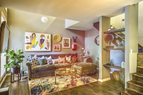 Cozy living room with a brown sofa, colorful artwork, plants, and a stylish staircase in a modern home.