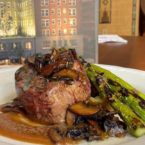 A plate of grilled steak topped with sautéed mushrooms and served with charred asparagus on a white dish.